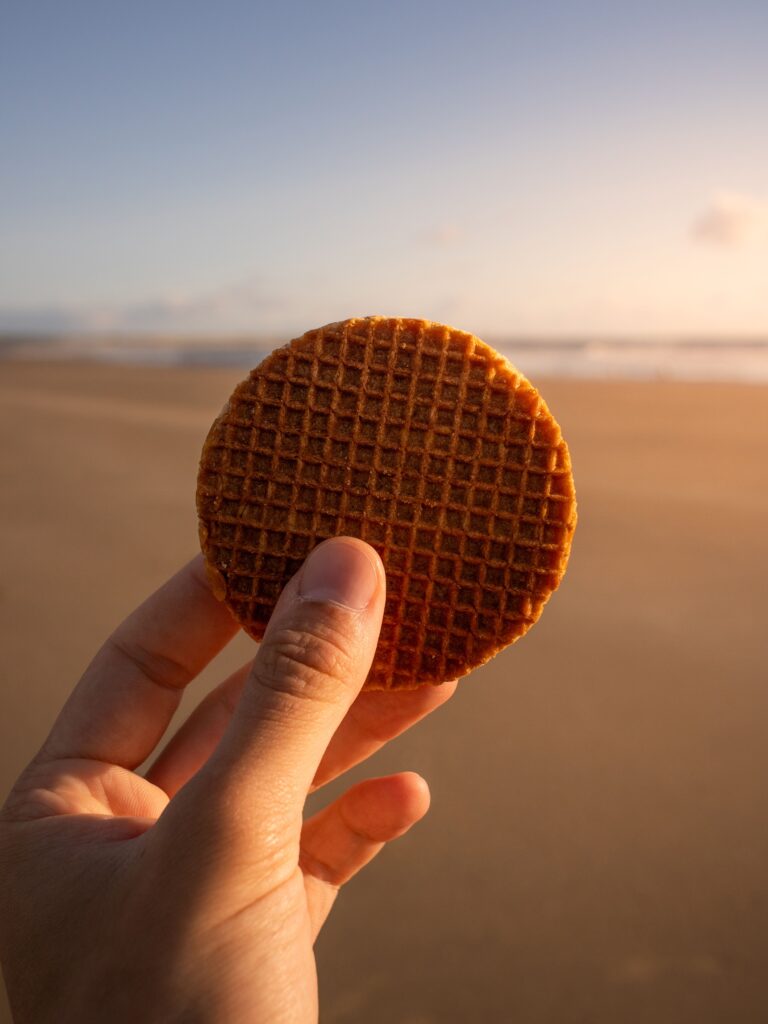 A fresh Dutch stroopwafel held up against a sunny North Sea beach background in the Netherlands.